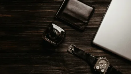 Flat lay of business essentials on dark wood: black leather wallet, glass camera, metal multi-dial wristwatch, and a silver laptop corner.](note: ensure proper punctuation)