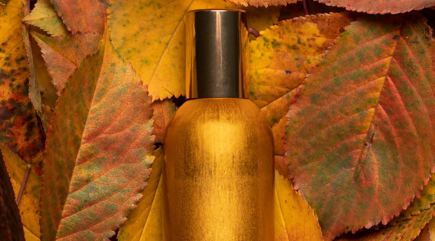 Hurmanski aromaty voseny vid tsukerni do molochnoho baru Gold perfume bottle with a black cap nestled among orange and yellow autumn leaves.