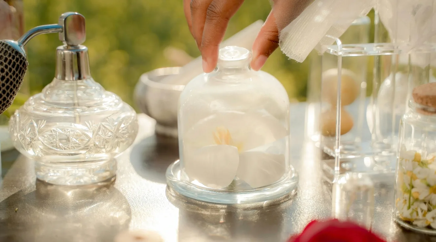 Parfumeriya za sezonamy shcho nosyty vzymku, vlitku ta mizh nymy Person places a white flower under a glass cloche on a reflective silver table, beside crystal perfume bottles and decor.