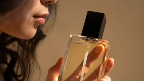 Parfumy dlya pobachennya, spivbesidy chy vazhlyvoyi zustrichi yak aromat vplyvaye na vrazhennya A person holding a clear glass perfume bottle with a black cap near their lips, as if about to apply scent.